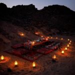Elegant dining table set up in the midst of a desert landscape with soft candlelight illuminating the scene.